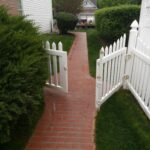 A white picket gate with a brick path. Greenery surrounds it.