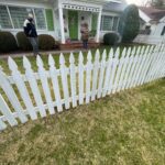 A white picket fence in front f a house with a green door and green shutters. Two people stand outside of it.